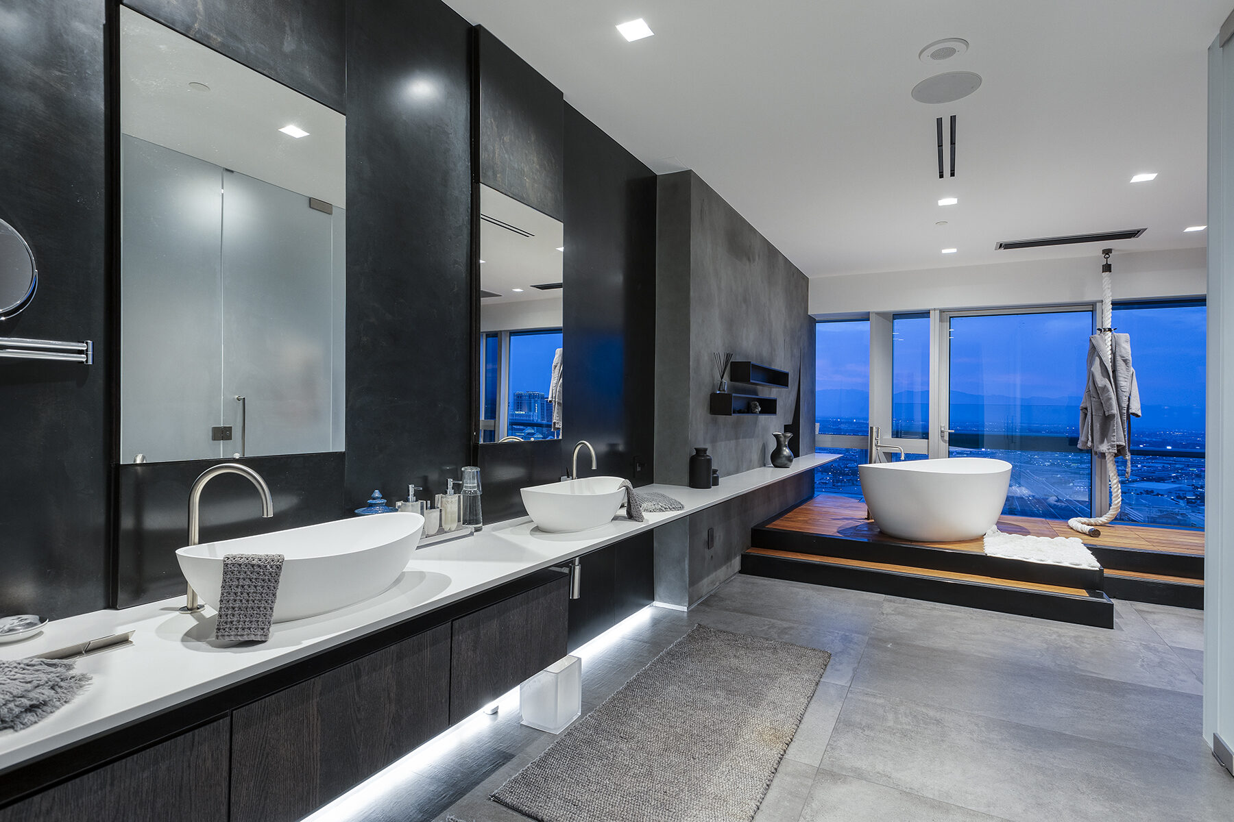 Penthouse master bath with a freestanding tub set against floor to ceiling windows overlooking the city at dusk