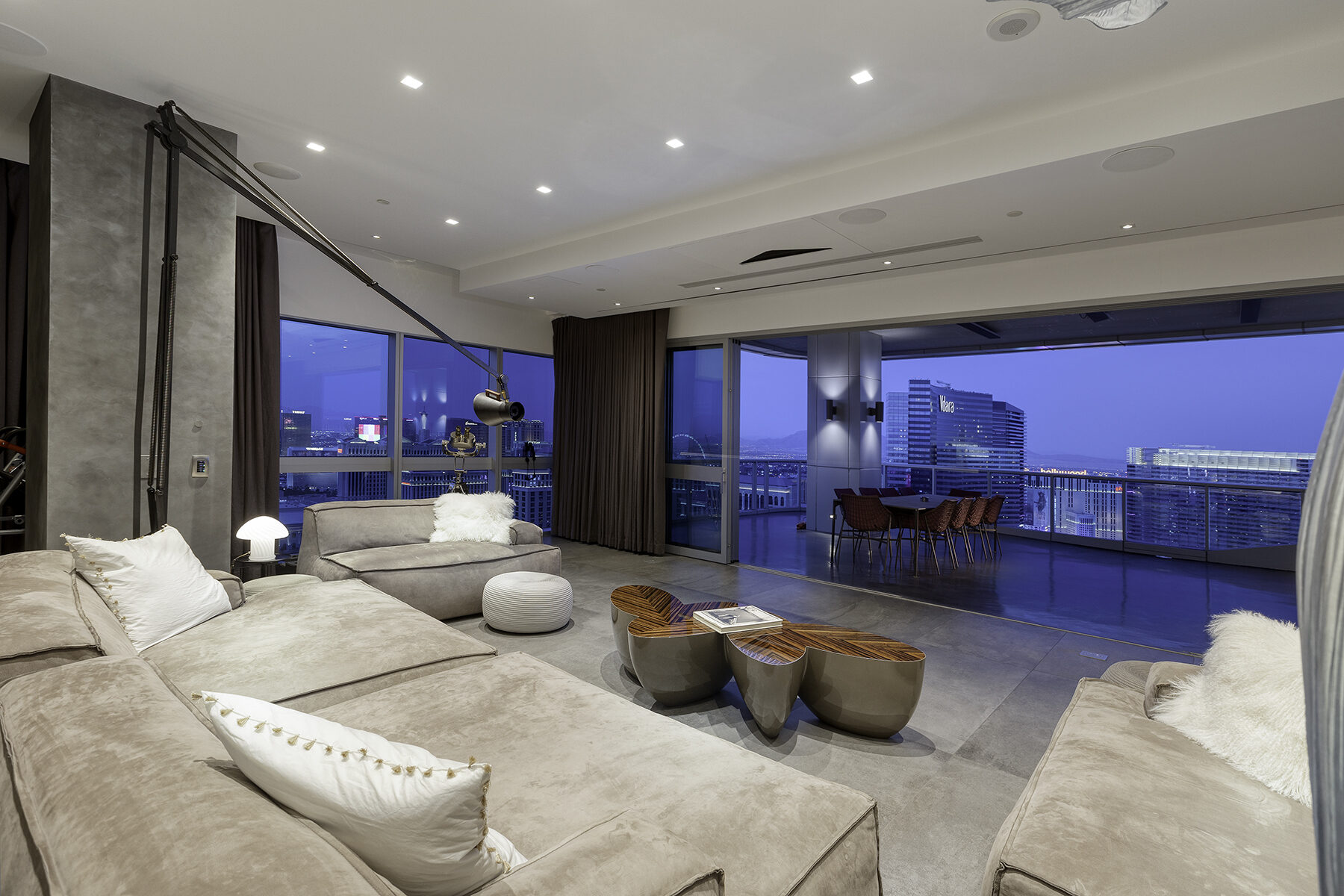 Penthouse great room at dusk with the pocket doors open and the drop-down screen retracted, the Las Vegas skyline unobstructed beyond the balcony